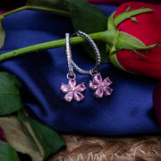 A pair of silver hoop earrings with pink sakura drop embellishments, displayed on a blue fabric surface with a red rose and leaves in the background.